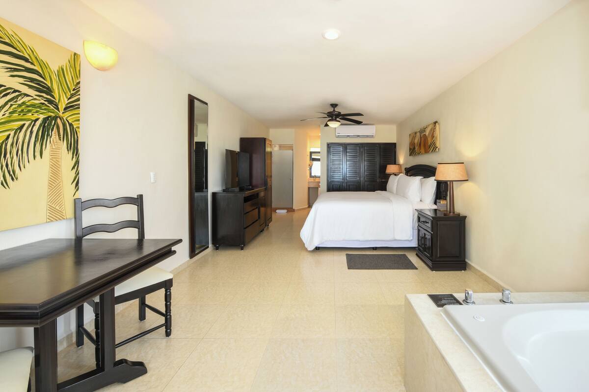 Habitación de hotel con cama, escritorio, silla, televisor, ventilador de techo, baño con bañera y un mural de palmeras.