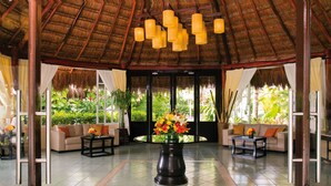 A spacious lobby with a thatched roof, comfortable seating, a central table with flowers, and large windows offering views of greenery.