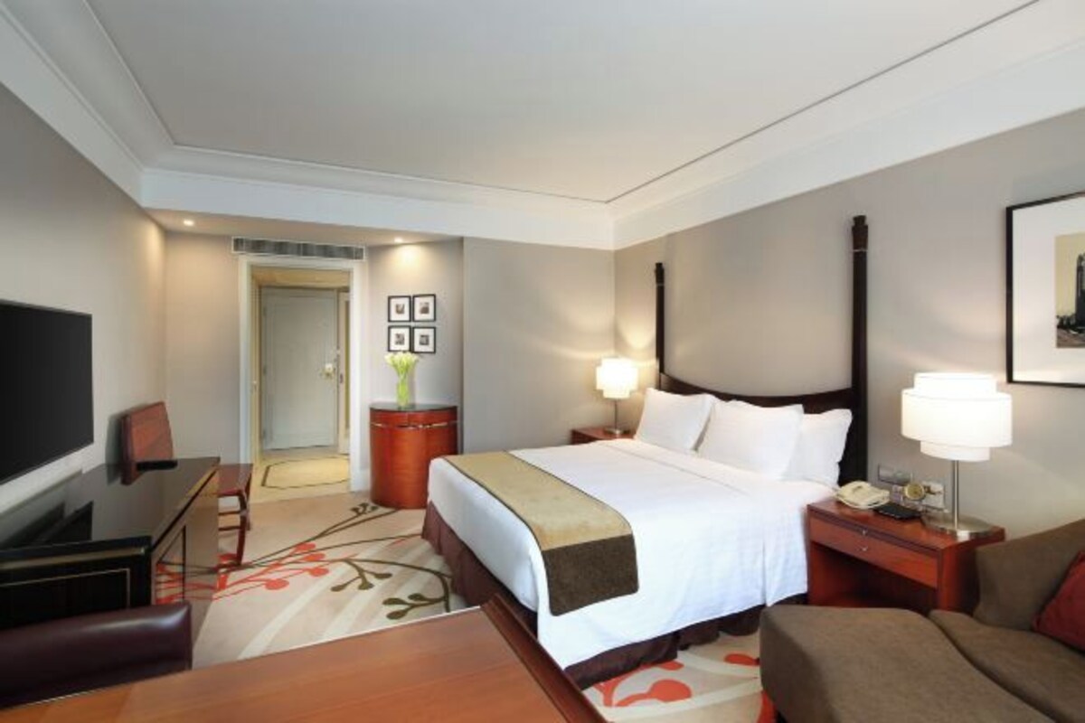 Quarto de hotel com cama, mesa de cabeceira, luminária, televisão, escrivaninha e cadeira.