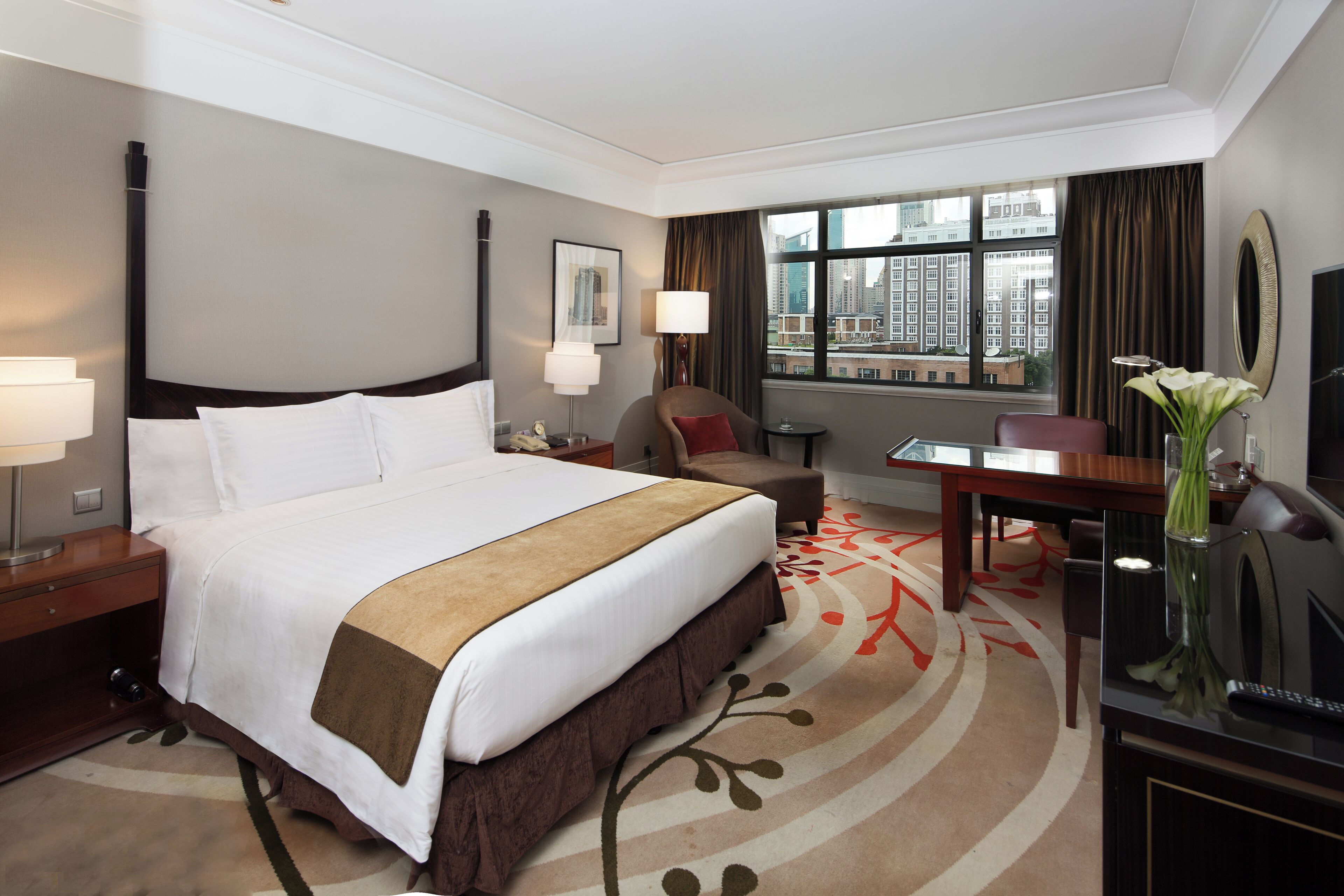 A hotel room with a large bed, a desk, a chair, a TV, and a window with a city view.