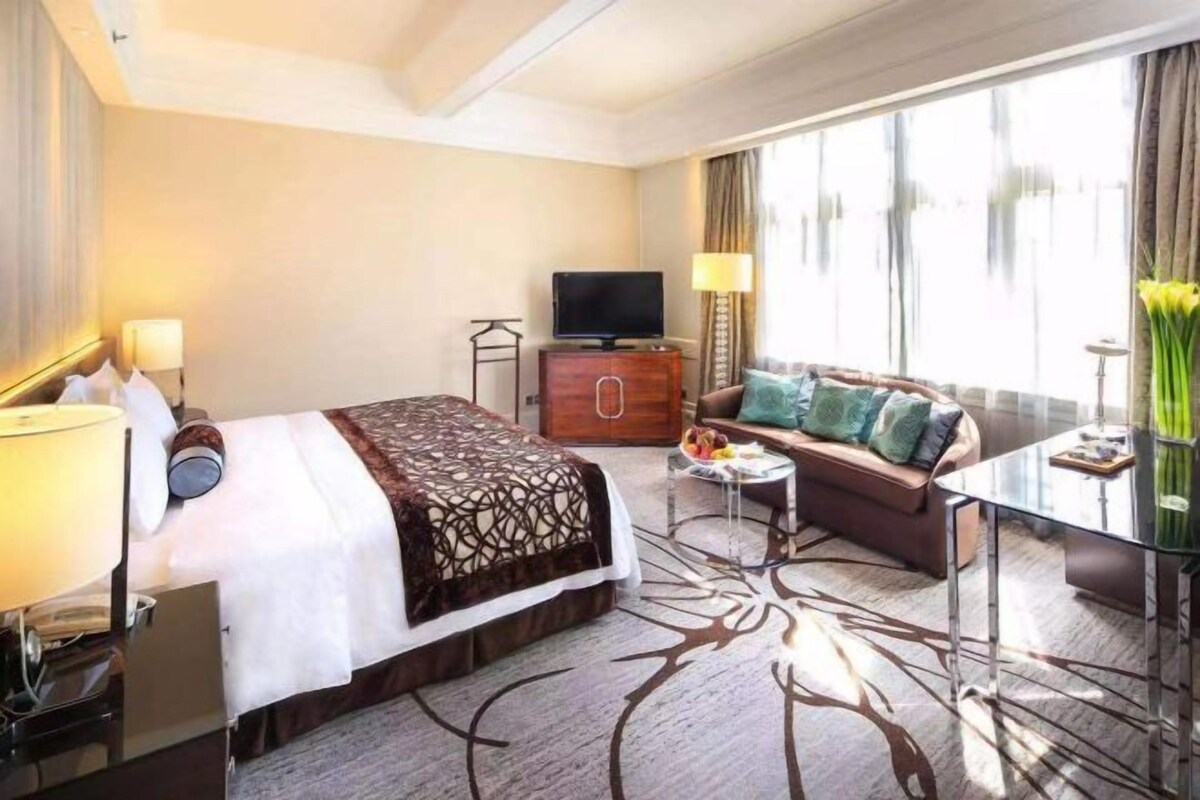 Habitación de hotel con cama, sofá, televisión, una mesita y ventana con cortinas.