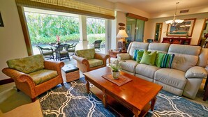 A living room with wicker furniture, a sofa with green and blue cushions, a wooden coffee table, and a patterned rug. - Villages at Mauna Lani (Kamuela)