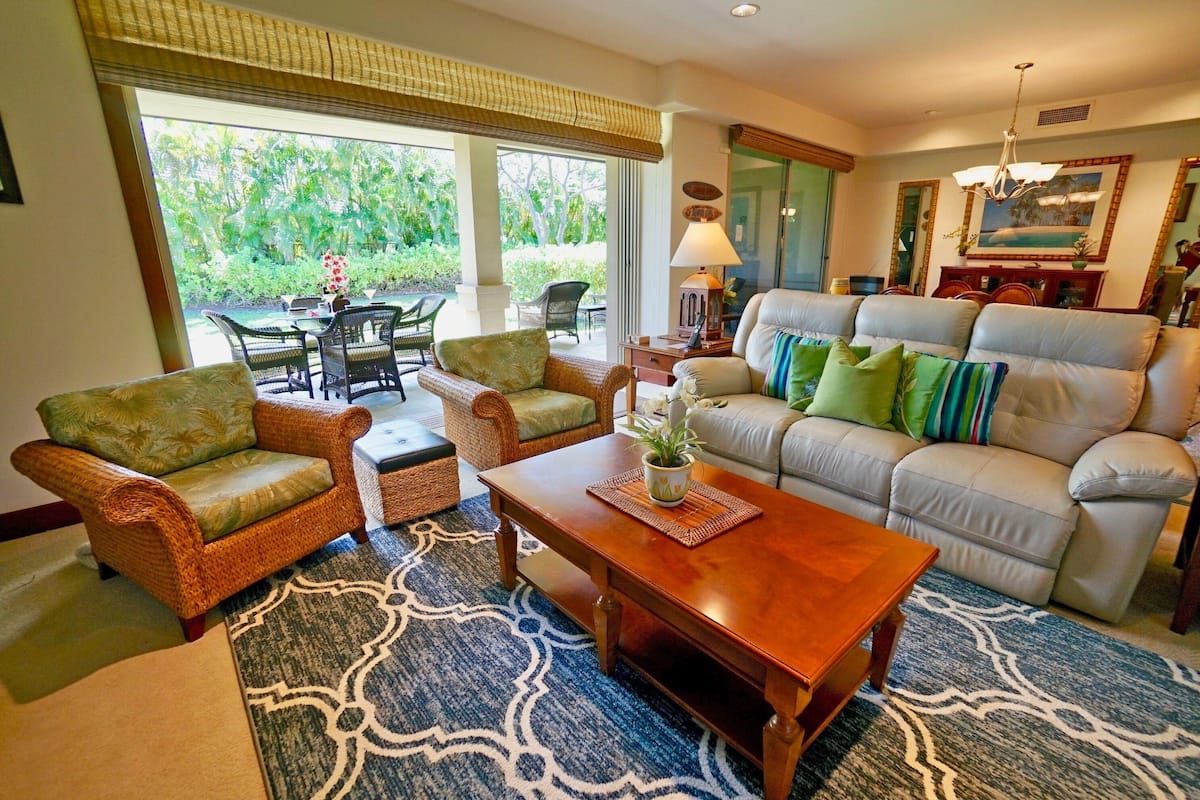 A living room with wicker furniture, a sofa with green and blue cushions, a wooden coffee table, and a patterned rug.