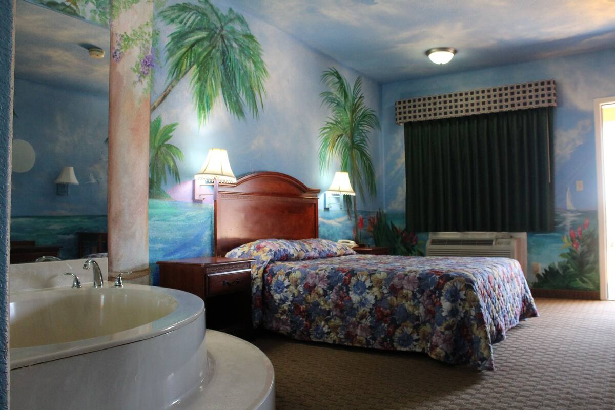 A hotel room with a large bathtub, a bed with floral bedding, and a mural of palm trees on the wall.