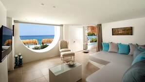 A modern living room with a large window offering a view of the sea and thatched umbrellas, a comfortable sofa, a glass coffee table, and a television. - Out Of The Blue All Inclusive Resort & Spa (Malevizi)