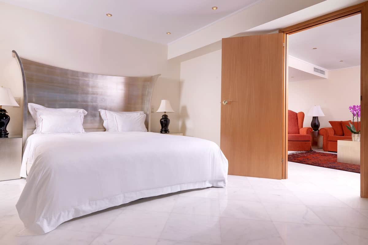 Une chambre spacieuse avec un grand lit, deux tables de chevet avec des lampes, une porte en bois donnant sur une autre pièce, et un canapé rouge dans le salon attenant.