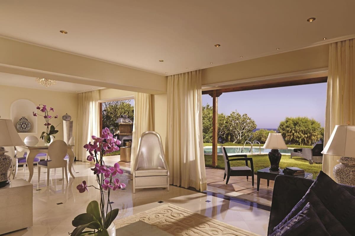 Un vaste salon avec vue sur une piscine et un espace verdoyant. On y trouve un coin repas avec une table et des chaises, ainsi qu’un coin salon avec un canapé et une lampe.