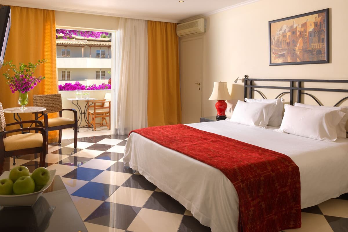Une chambre d’hôtel avec un sol à carreaux, un lit avec du linge de maison blanc et une couverture rouge, une table de chevet avec deux lampes, une table avec des chaises et un balcon orné de fleurs violettes.