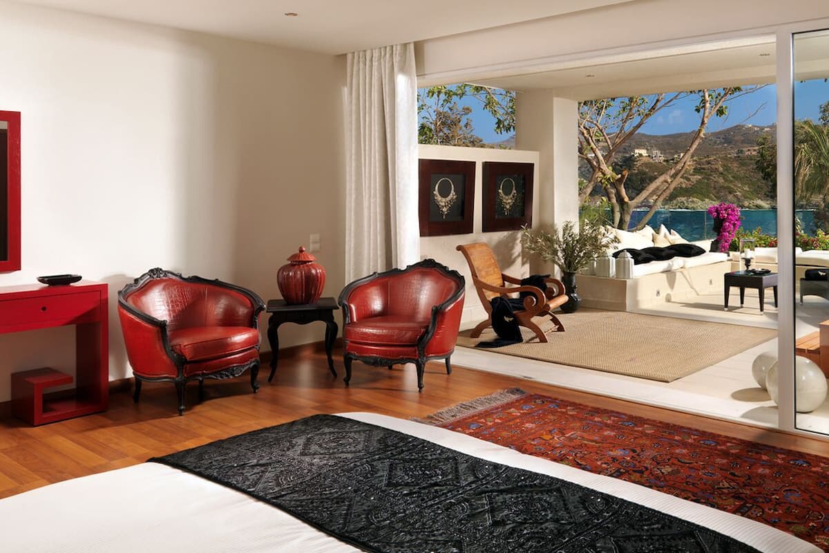 Une chambre moderne avec un grand lit, un fauteuil en cuir rouge, un bureau en bois et une vue sur un jacuzzi et un espace extérieur offrant un paysage pittoresque.