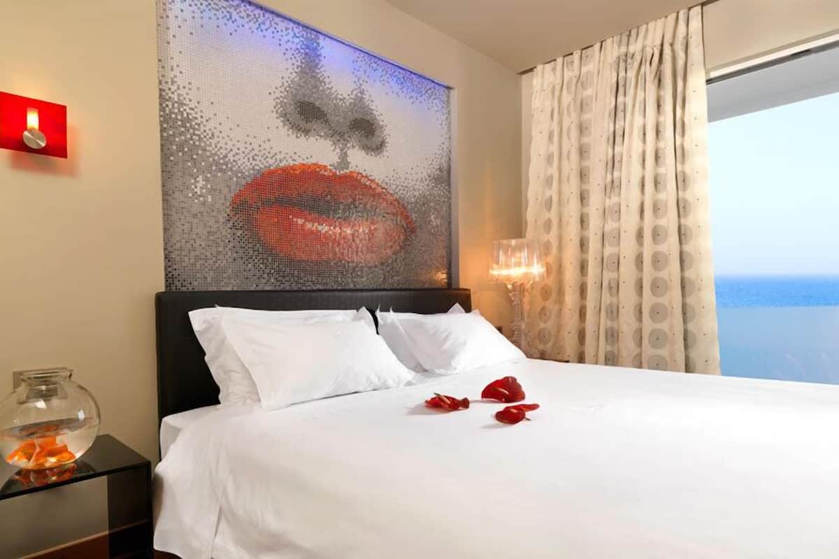 Une chambre d’hôtel avec un lit, une grande œuvre d’art représentant des lèvres, une table de chevet avec un bocal à poissons et une vue sur la mer par la fenêtre.