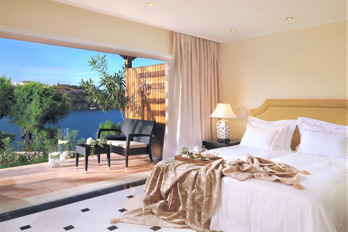 Une chambre d’hôtel avec un lit, une chaise, une table avec un plateau, une lampe et une vue sur la mer.