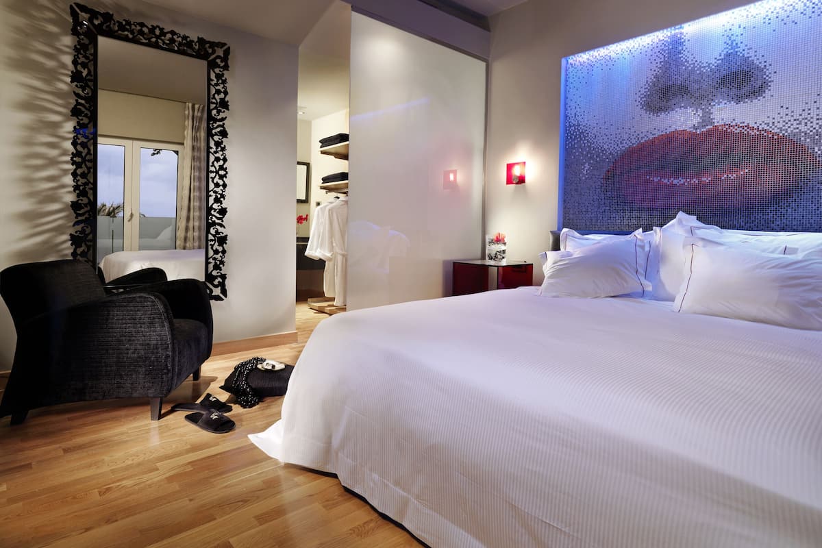 Une chambre d’hôtel moderne dotée d’un grand lit, d’un coin salon avec un fauteuil, d’une armoire et d’une grande fenêtre offrant une vue dégagée.