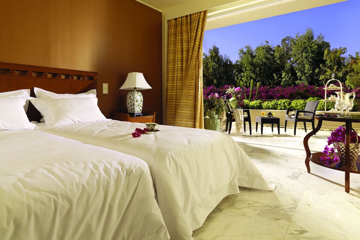 Une chambre d’hôtel avec deux lits, une table de chevet, une lampe et une vue sur un jardin fleuri, avec une table à l’extérieur.