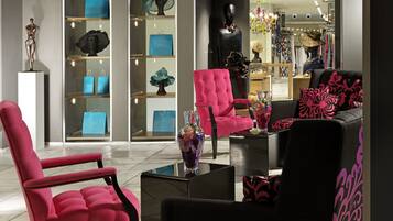 A modern interior with a seating area featuring a pink armchair and a black sofa with pink floral cushions. There are glass display cases with blue shopping bags and decorative items.