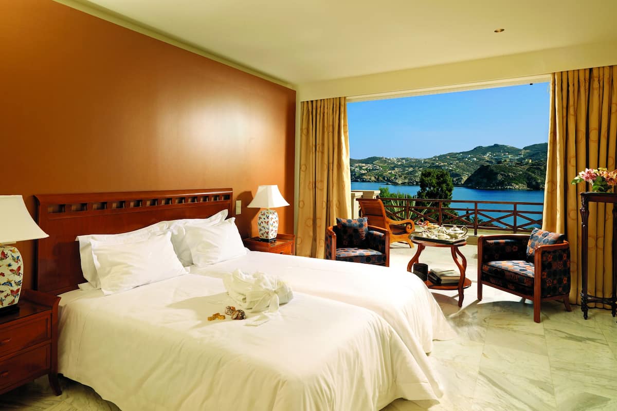 Une chambre d’hôtel avec un grand lit, deux tables de chevet avec des lampes, une petite table avec des chaises et un balcon offrant une vue sur la mer et les collines.