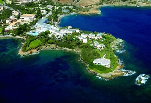 Vista aérea de un complejo turístico frente al mar, con varias construcciones, una playa y aguas azules cristalinas.
