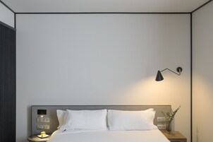 A modern hotel room with a neatly made bed, a bedside table with a lamp, and a wall-mounted control panel.