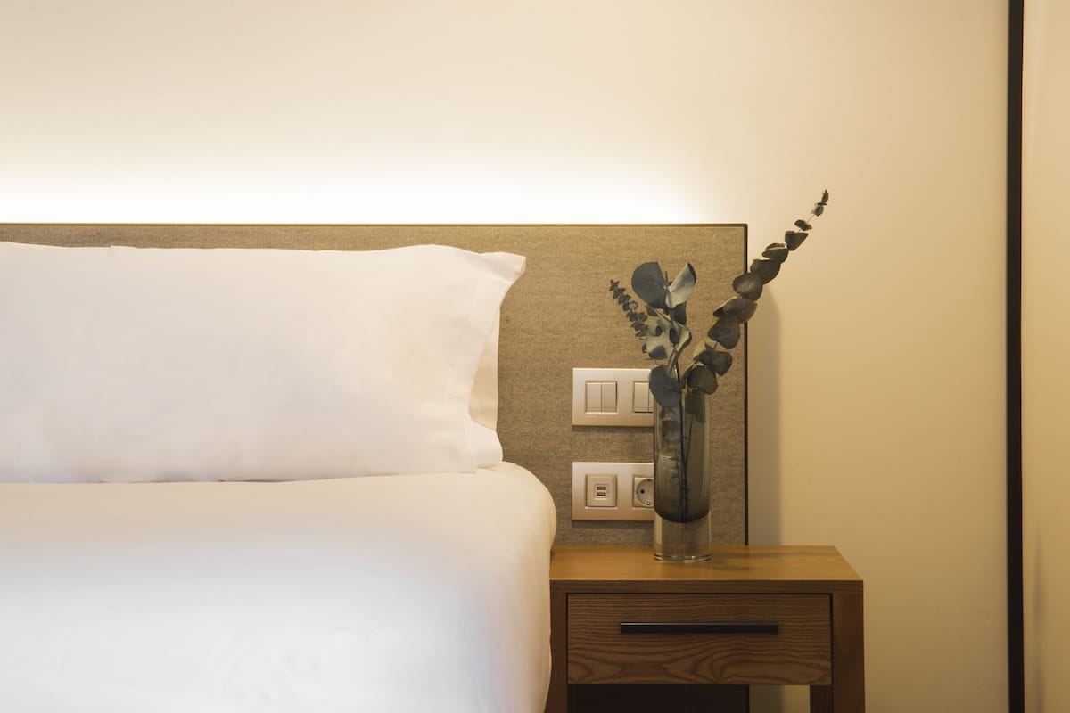 Una cama bien hecha con ropa blanca, una mesita de noche con un jarrón de plantas verdes, y un interruptor de luz en la pared.