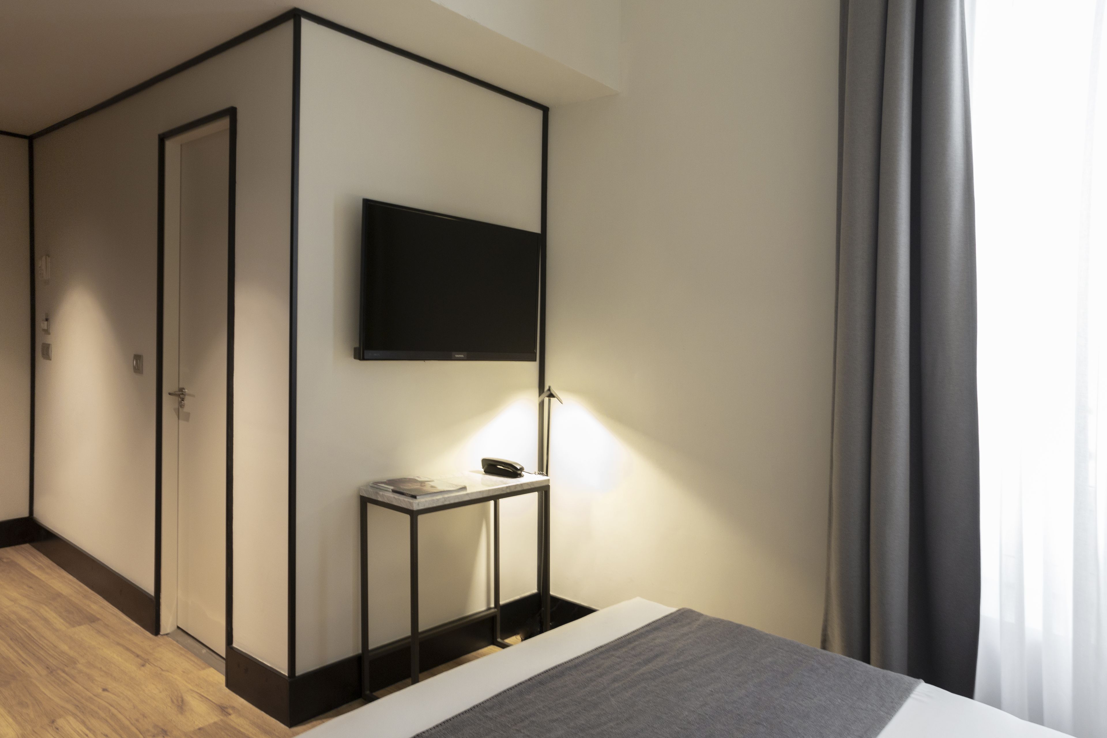 Una habitación de hotel moderna con un televisor de pantalla plana montado en la pared, una mesita de noche con lámpara y suelo de madera.
