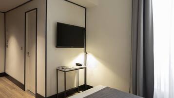 Una habitación de hotel moderna con un televisor de pantalla plana montado en la pared, una mesita de noche con lámpara y suelo de madera.
