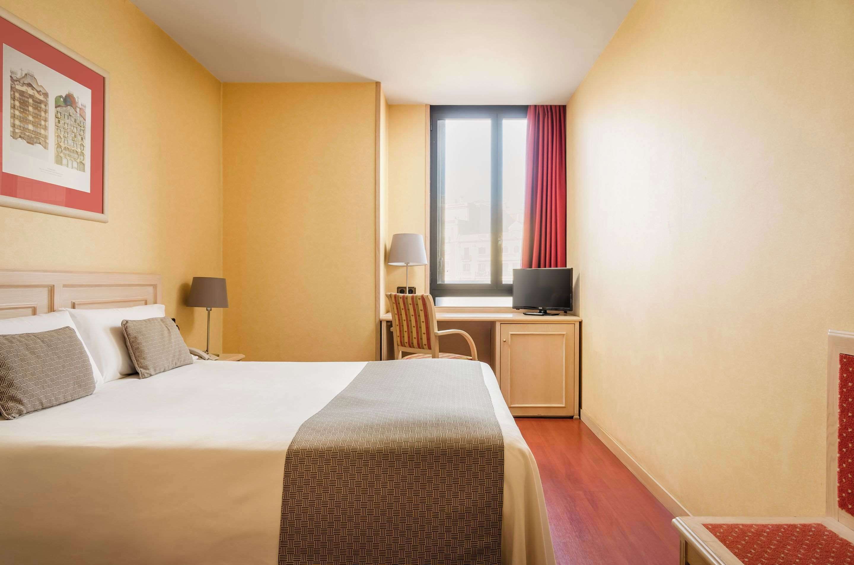 Ett hotellrum med en säng, ett skrivbord med en dator, en stol, en lampa och ett fönster med gardiner.
