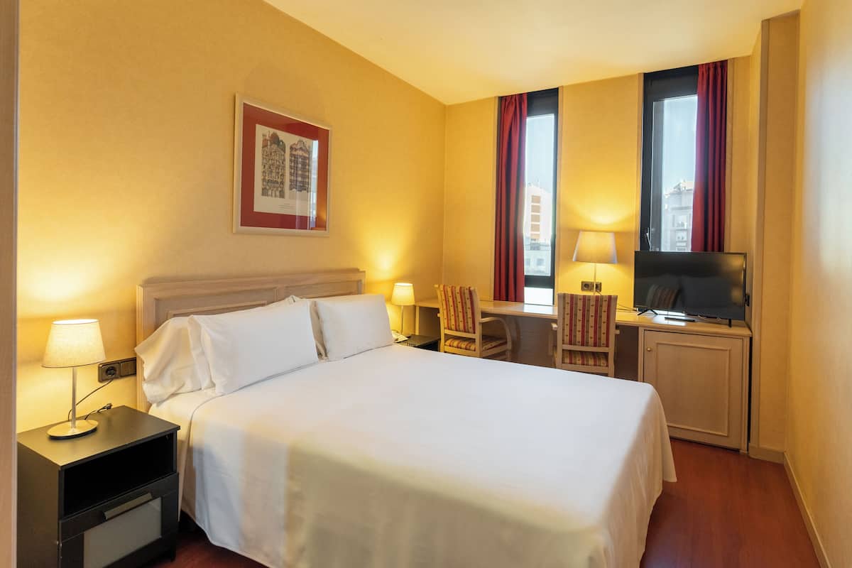 A hotel room with a bed, bedside tables, a desk with a chair, a television, and a window with curtains.