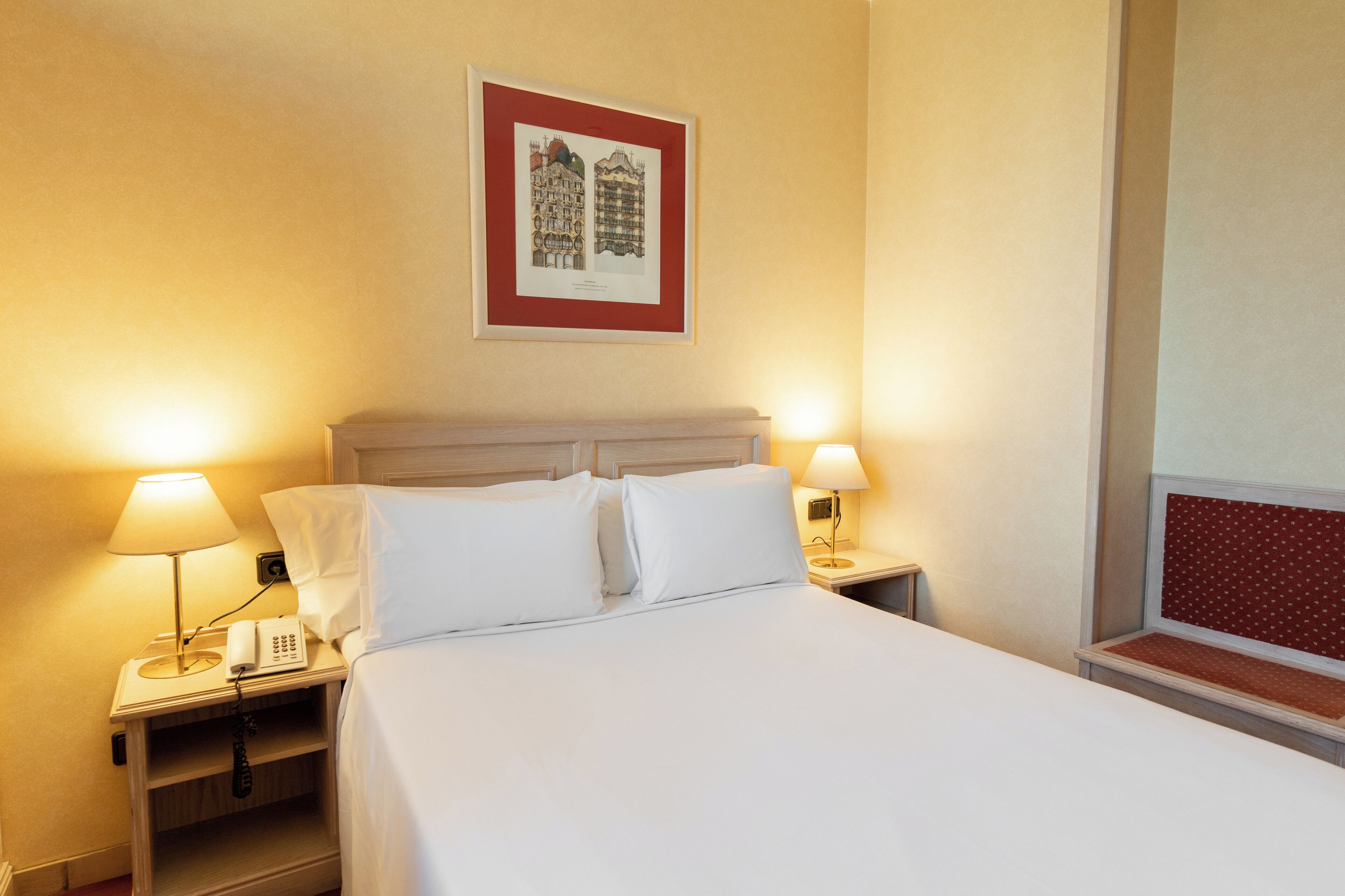 A neatly made bed with white linens, two bedside lamps, a framed picture on the wall, and a window with a red frame.
