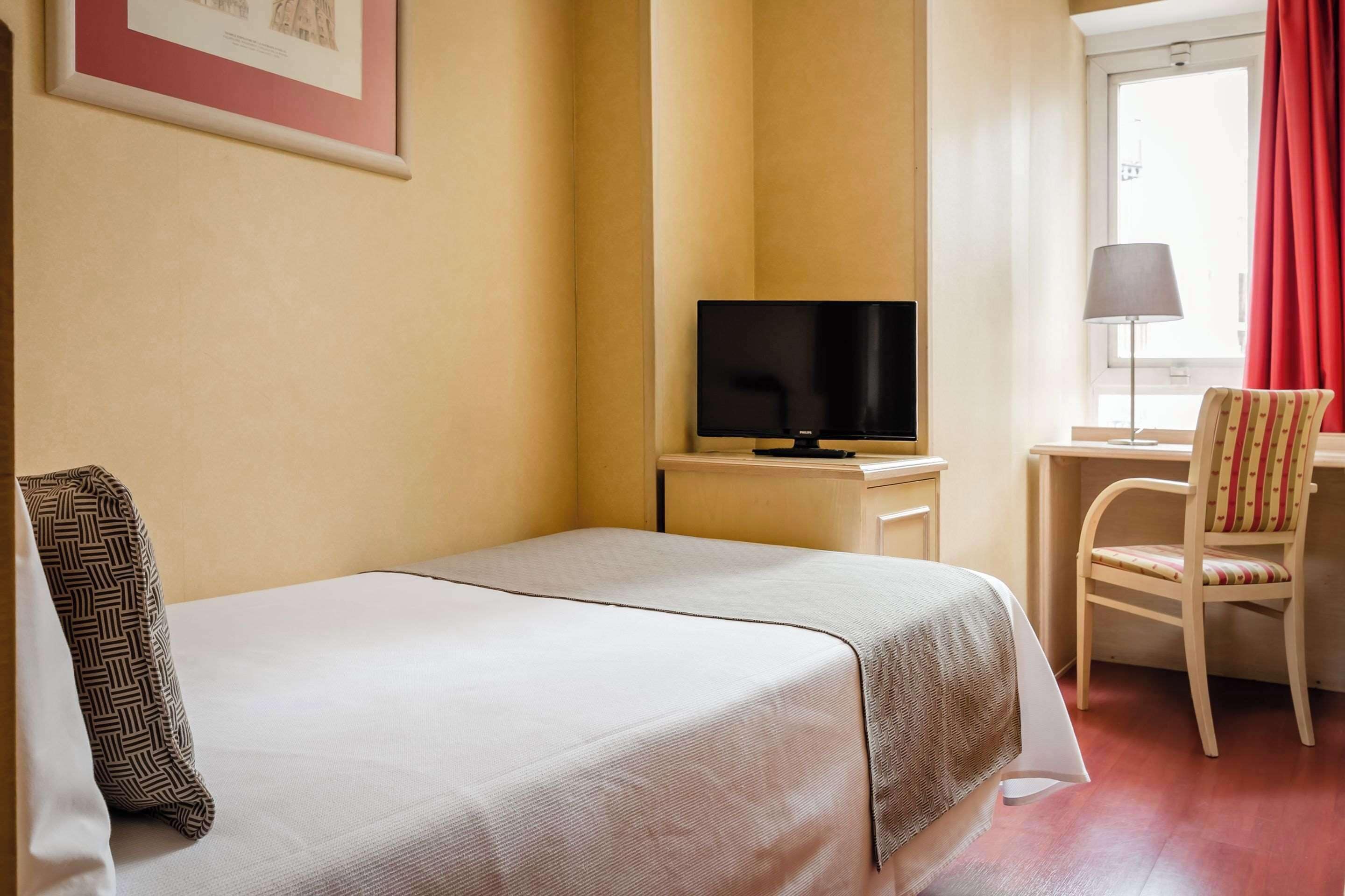 Una habitación de hotel con cama, escritorio con televisor, silla y ventana con cortinas rojas.