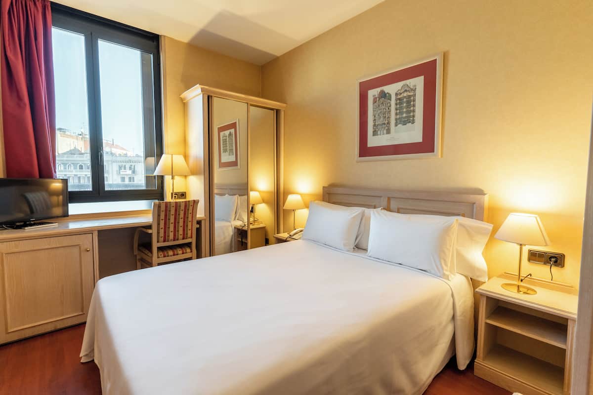 A hotel room with a bed, bedside tables, a desk with a chair, a television, and a window with curtains.