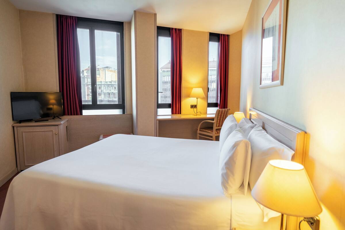 A hotel room with a large bed, a desk, a chair, a television, and two windows with curtains.