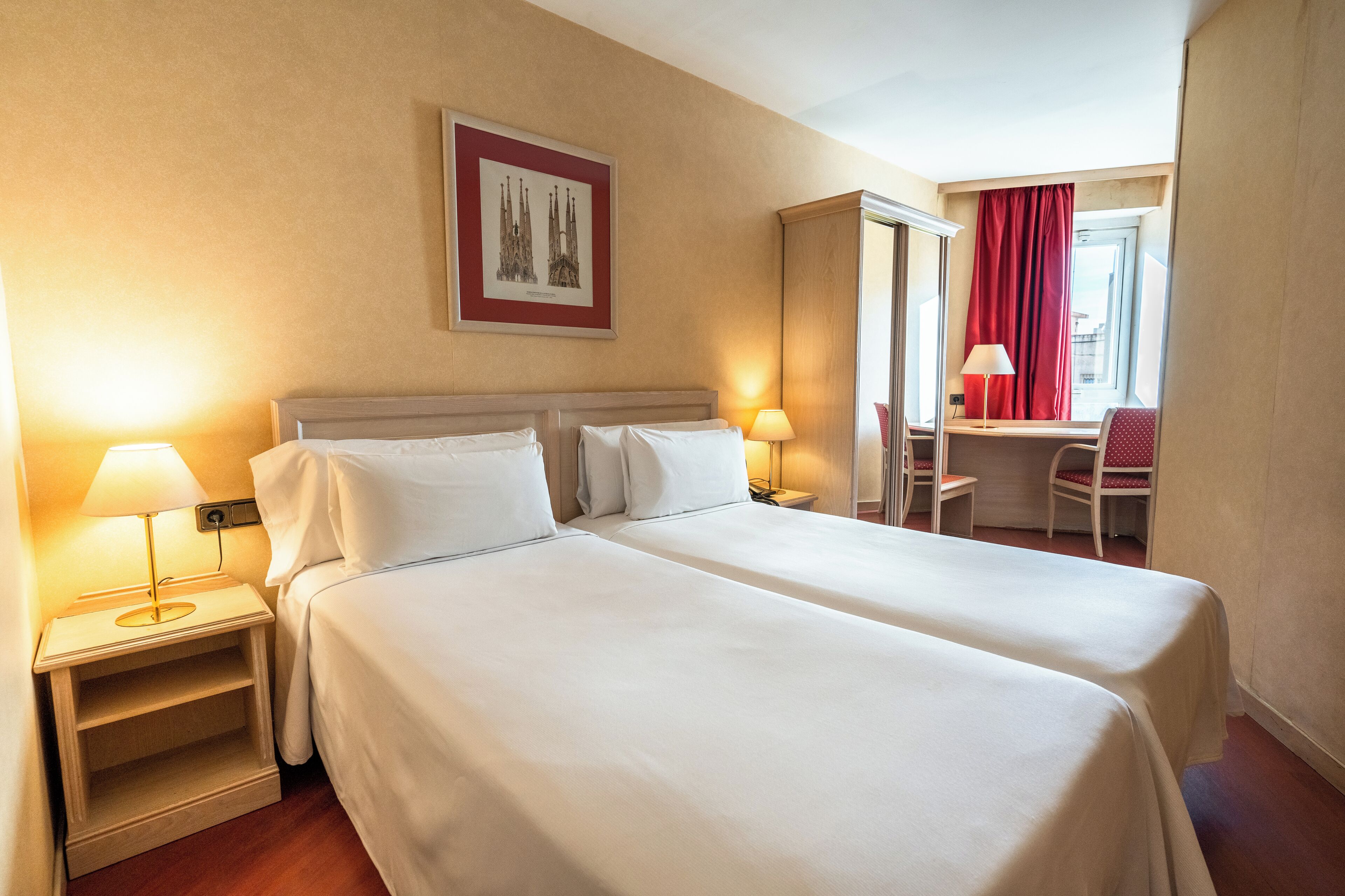 Habitación de hotel con dos camas, un escritorio, una silla y una ventana con cortinas rojas.
