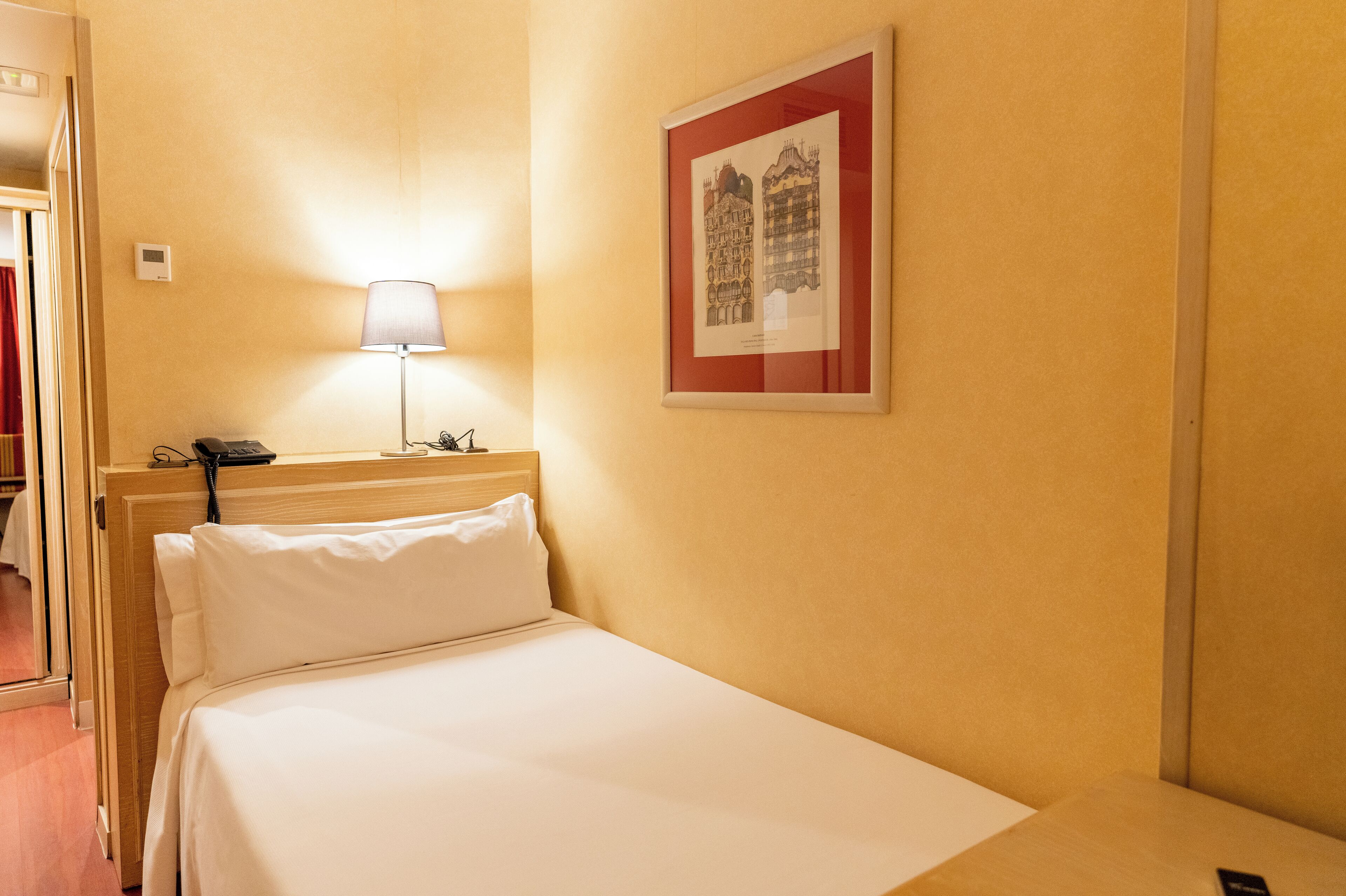 A hotel room with a bed, a bedside table, a lamp, a phone, and a framed picture on the wall.