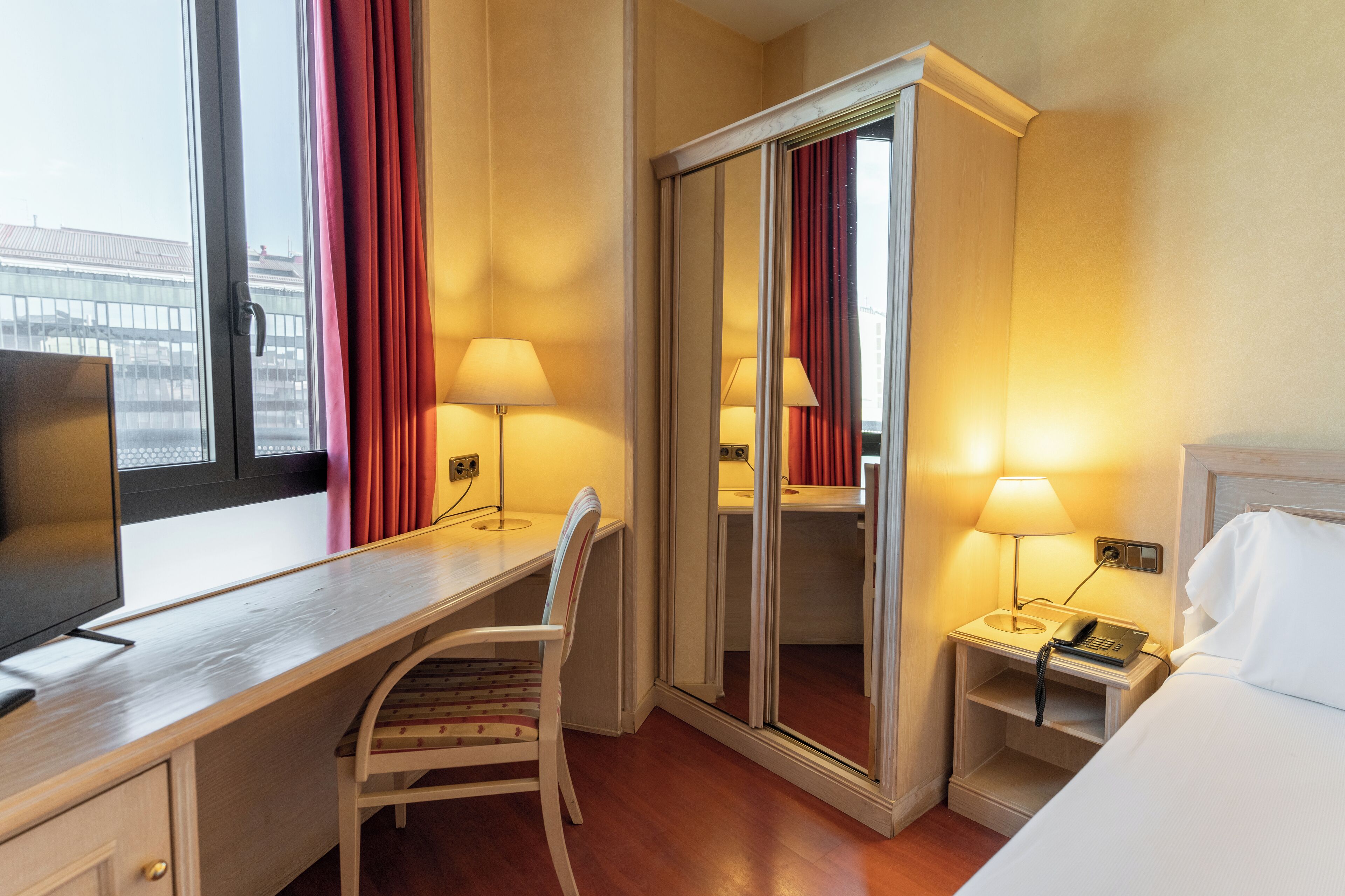 A hotel room with a desk, chair, bed, bedside table with lamps, and a mirror.