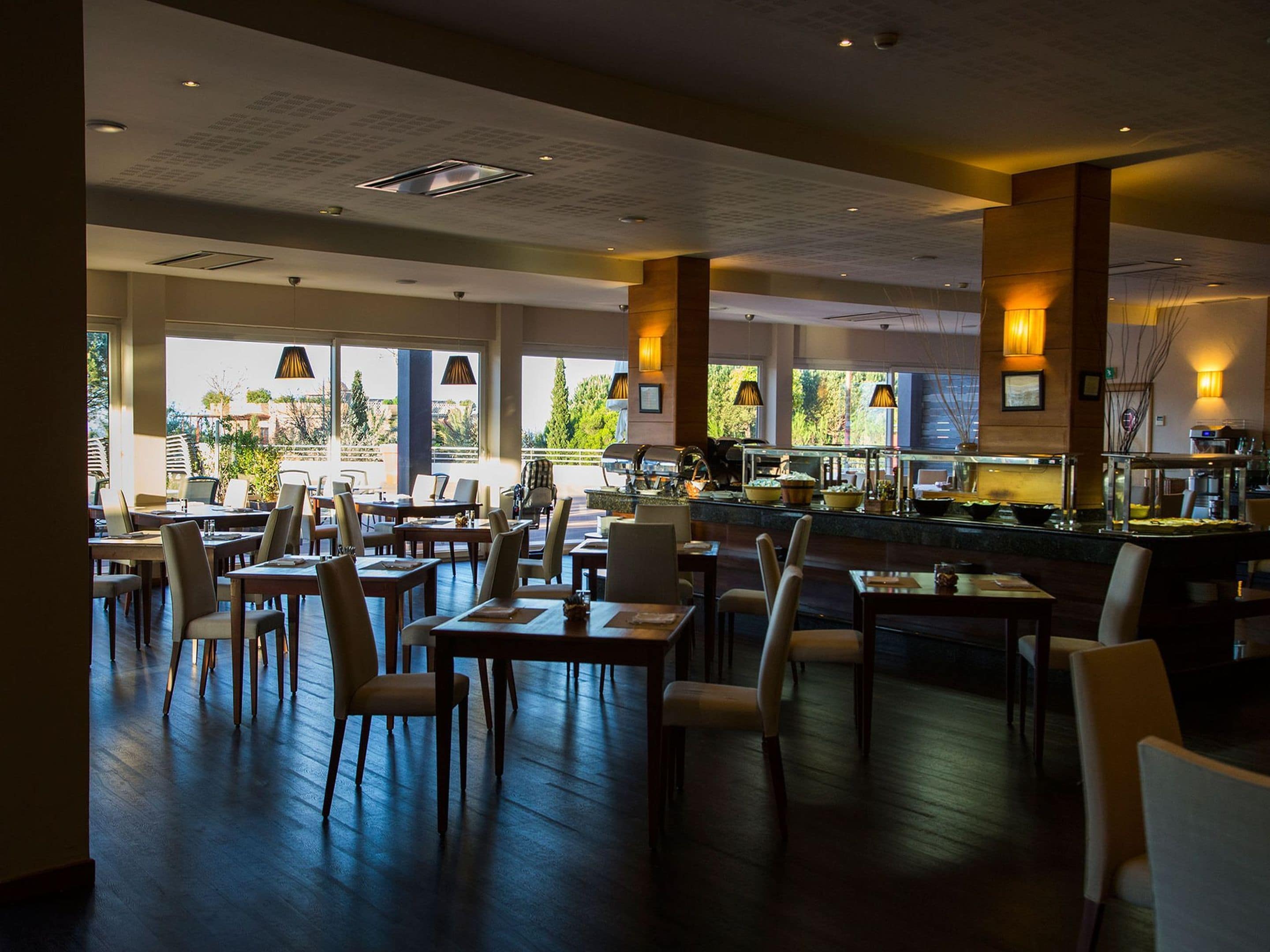A spacious restaurant with multiple dining tables and chairs, large windows offering views of the outdoors, and a buffet area with various food options.