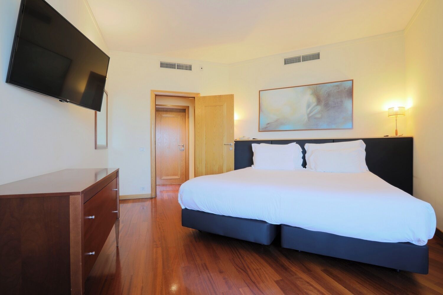 A hotel room with a bed, a TV mounted on the wall, a wooden dresser, and a painting on the wall.