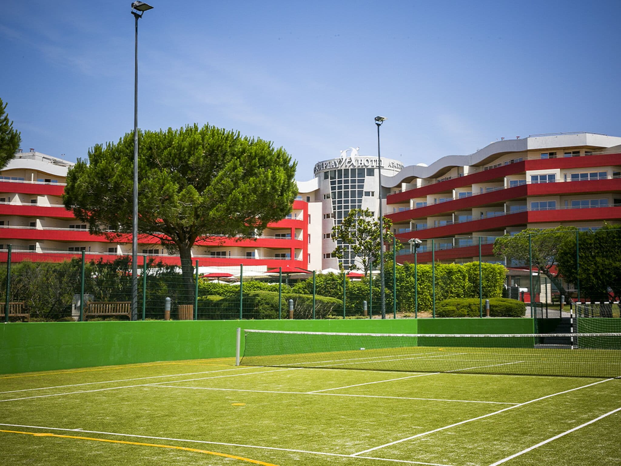 Ein Tennisplatz mit Netz, umgeben von grünem Zaun und Bäumen. Im Hintergrund ein Hotel mit roten und weißen Gebäuden und Balkonen.