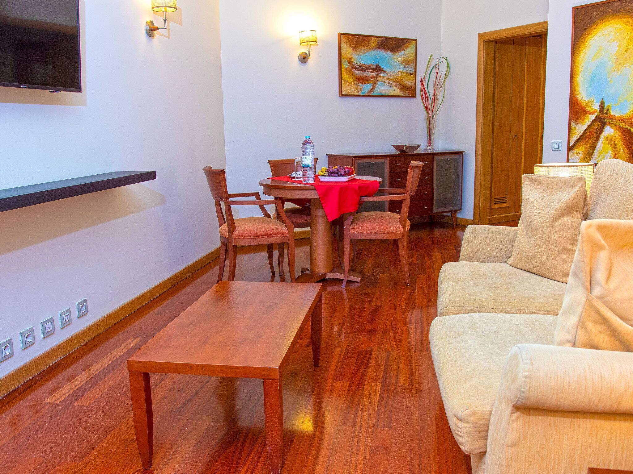 A hotel room with a wooden dining table, two chairs, a sofa, a flat-screen TV mounted on the wall, and artwork on the walls.