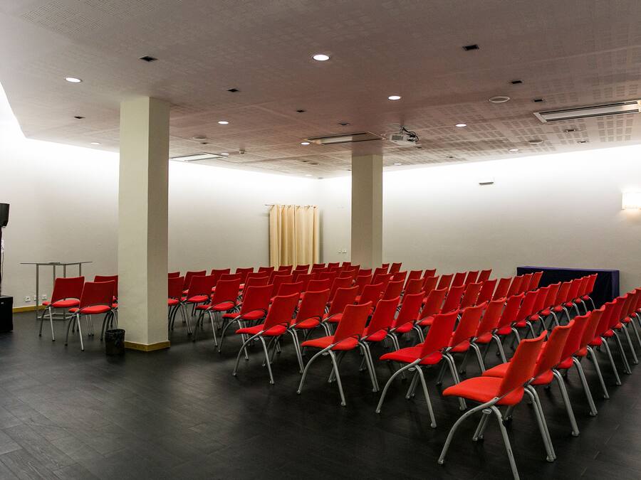 A conference room with rows of red chairs, a podium, and a projector screen.