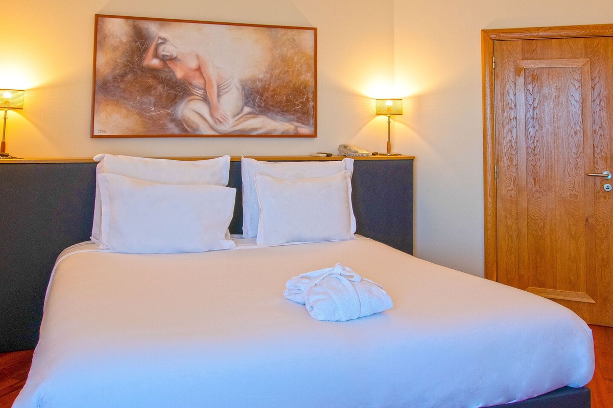Un lit bien fait, avec des draps blancs, deux oreillers et une serviette pliée. Il y a un tableau accroché au mur au-dessus du lit et une porte en bois sur la droite.