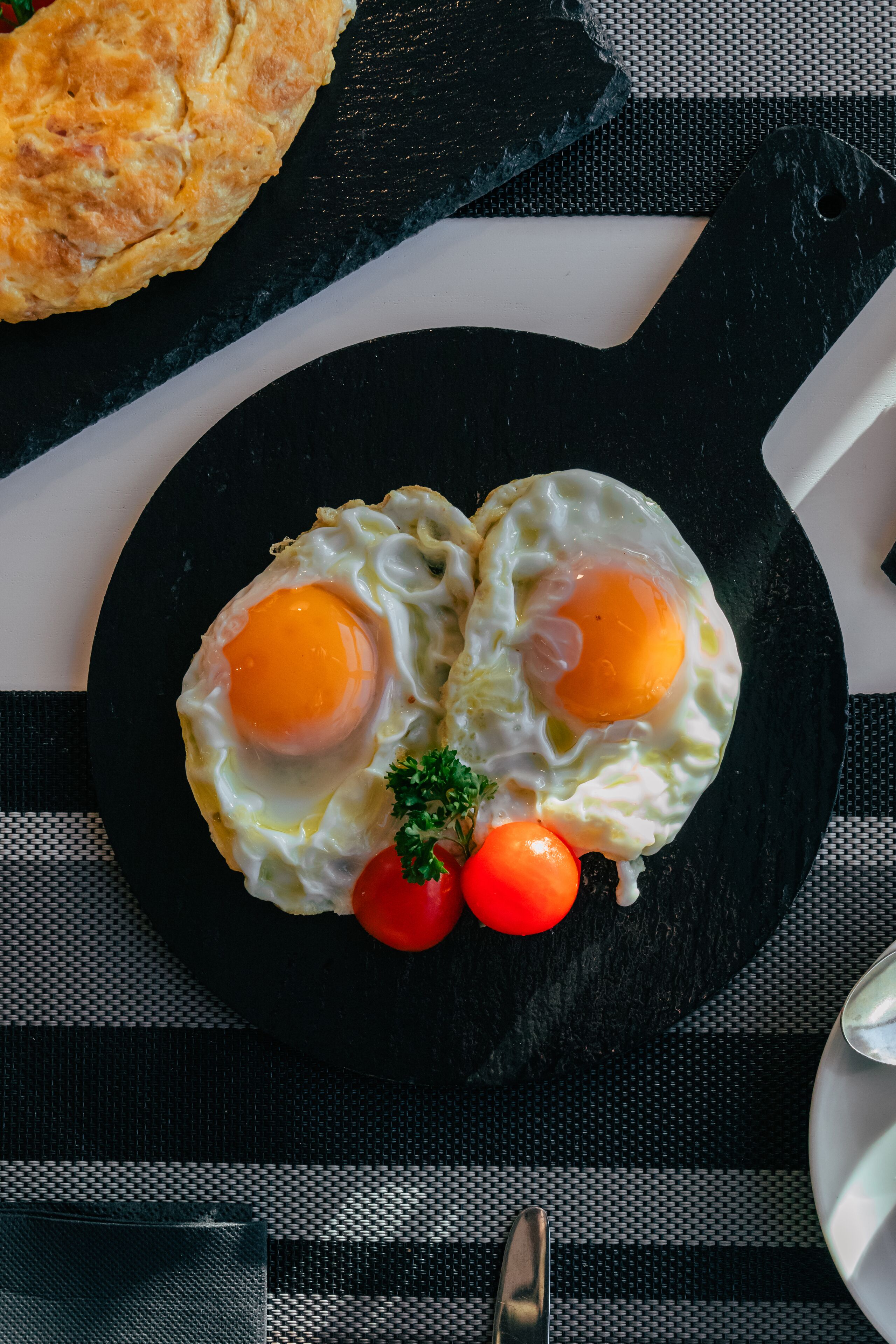 Una sartén negra sobre un mantel a rayas blancas y negras, con dos huevos fritos y tomates cherry encima.