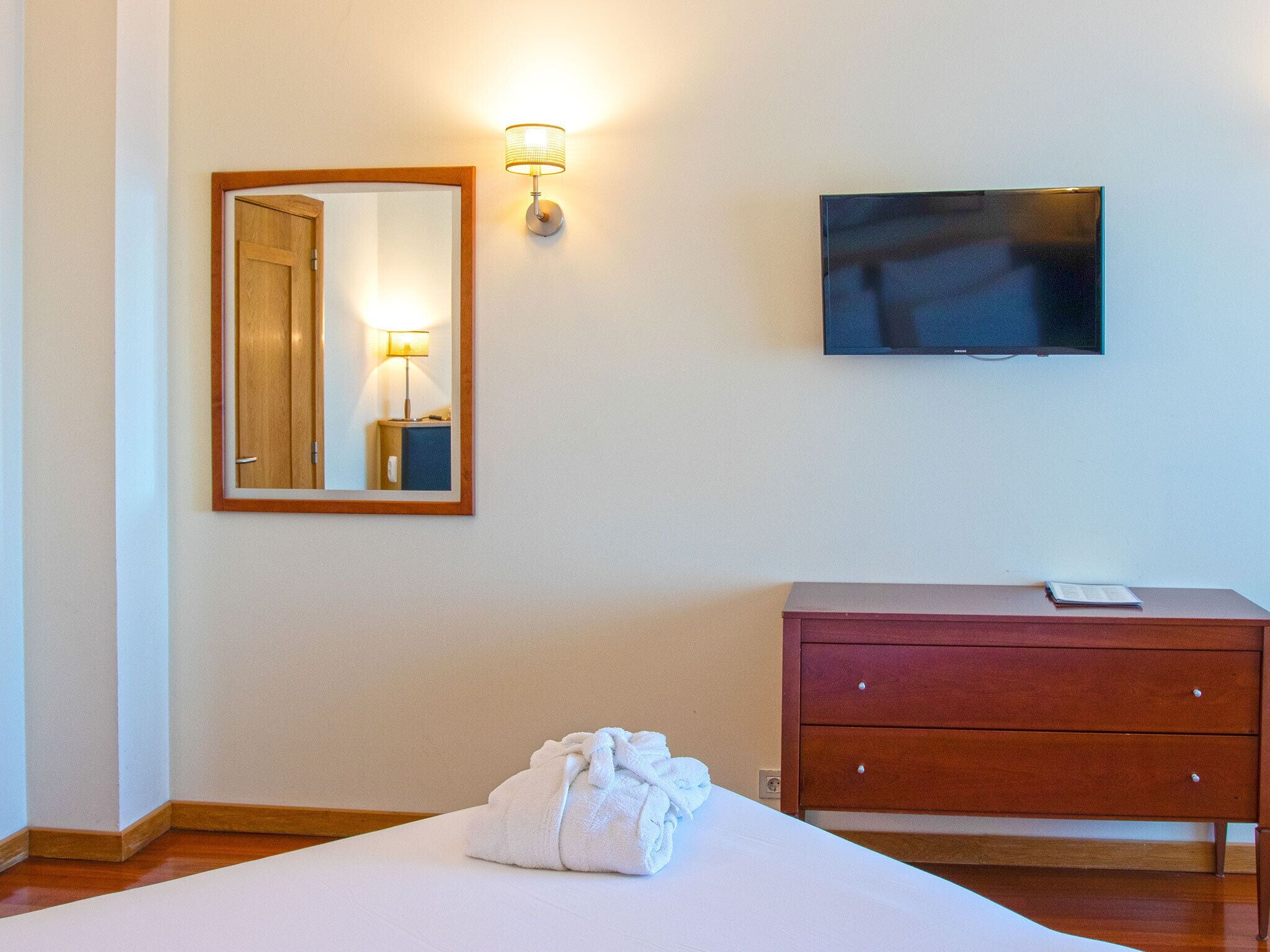 A hotel room with a bed, a wooden dresser, a television mounted on the wall, and a mirror with a lamp.