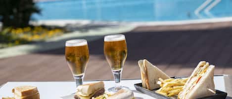 Una mesa con sándwiches, papas fritas y dos vasos de cerveza, con una piscina al fondo.