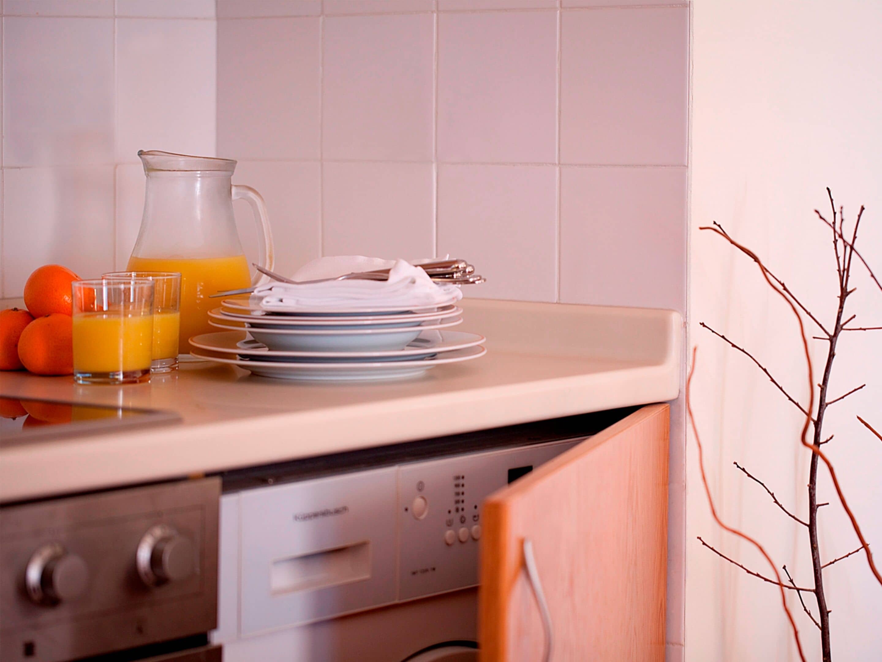 Une cuisine avec un plan de travail, une carafe de jus d’orange, une pile d’assiettes et quelques oranges.