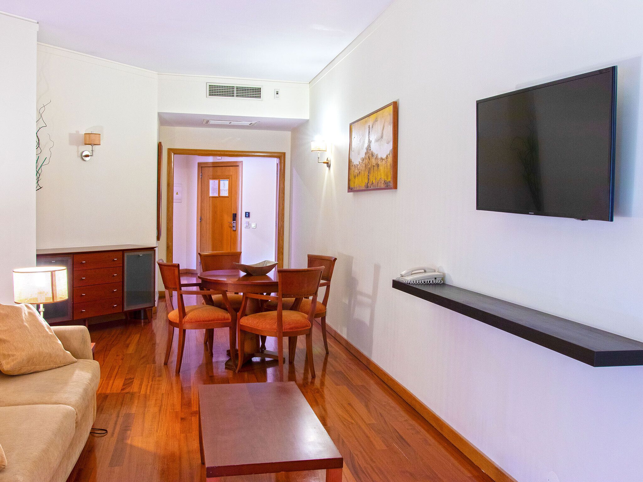 Una sala de estar con un sofá, una mesa de centro, una mesa de comedor con sillas, un televisor montado en la pared y un aparador.