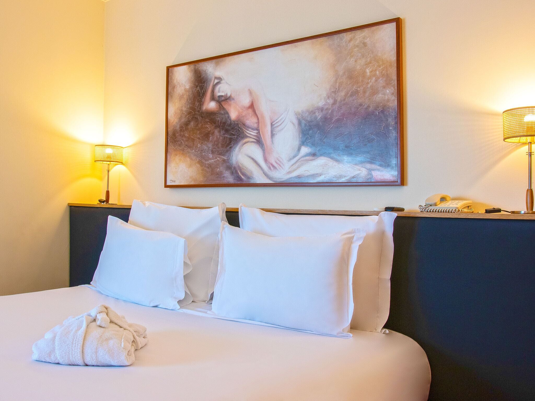 A hotel room with a bed, two bedside lamps, a painting on the wall, and a telephone on the nightstand.
