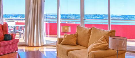 Una sala de estar con un sofá, una mesa y una silla. Hay una ventana con vista a un cuerpo de agua y un edificio rojo.
