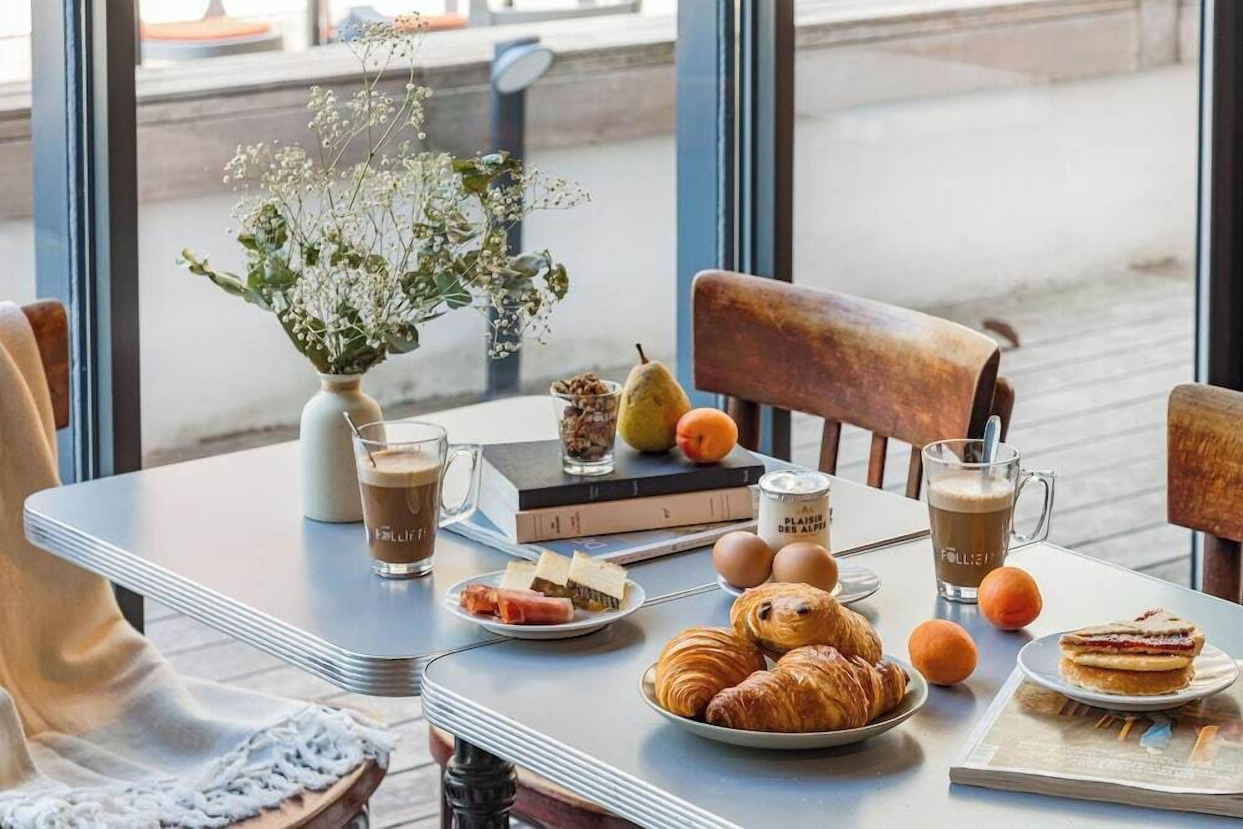 A table set with pastries, fruits, and drinks, a vase with flowers, and a book.