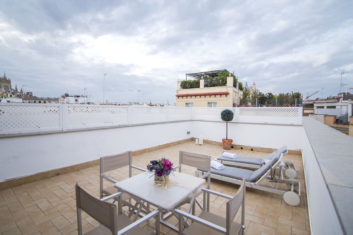 A rooftop terrace with a table and chairs, a lounge area with sun loungers, and a view of the cityscape.