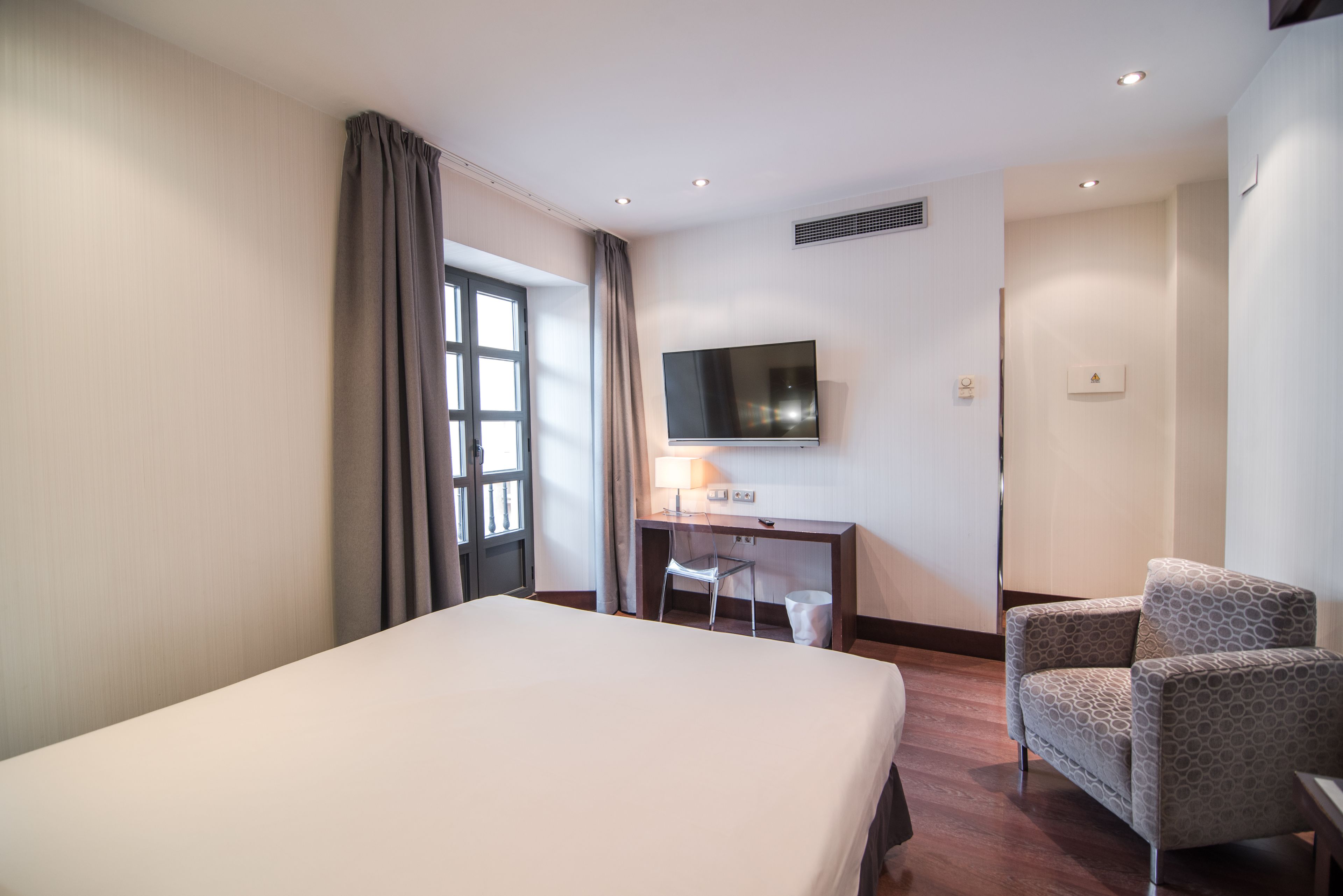 Habitación de hotel con una cama, un escritorio con silla, un televisor y una ventana con cortinas.