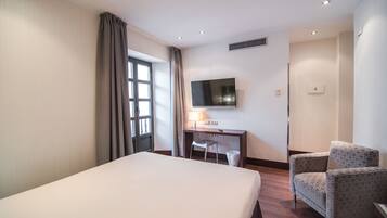 Habitación de hotel con una cama, un escritorio con silla, un televisor y una ventana con cortinas.